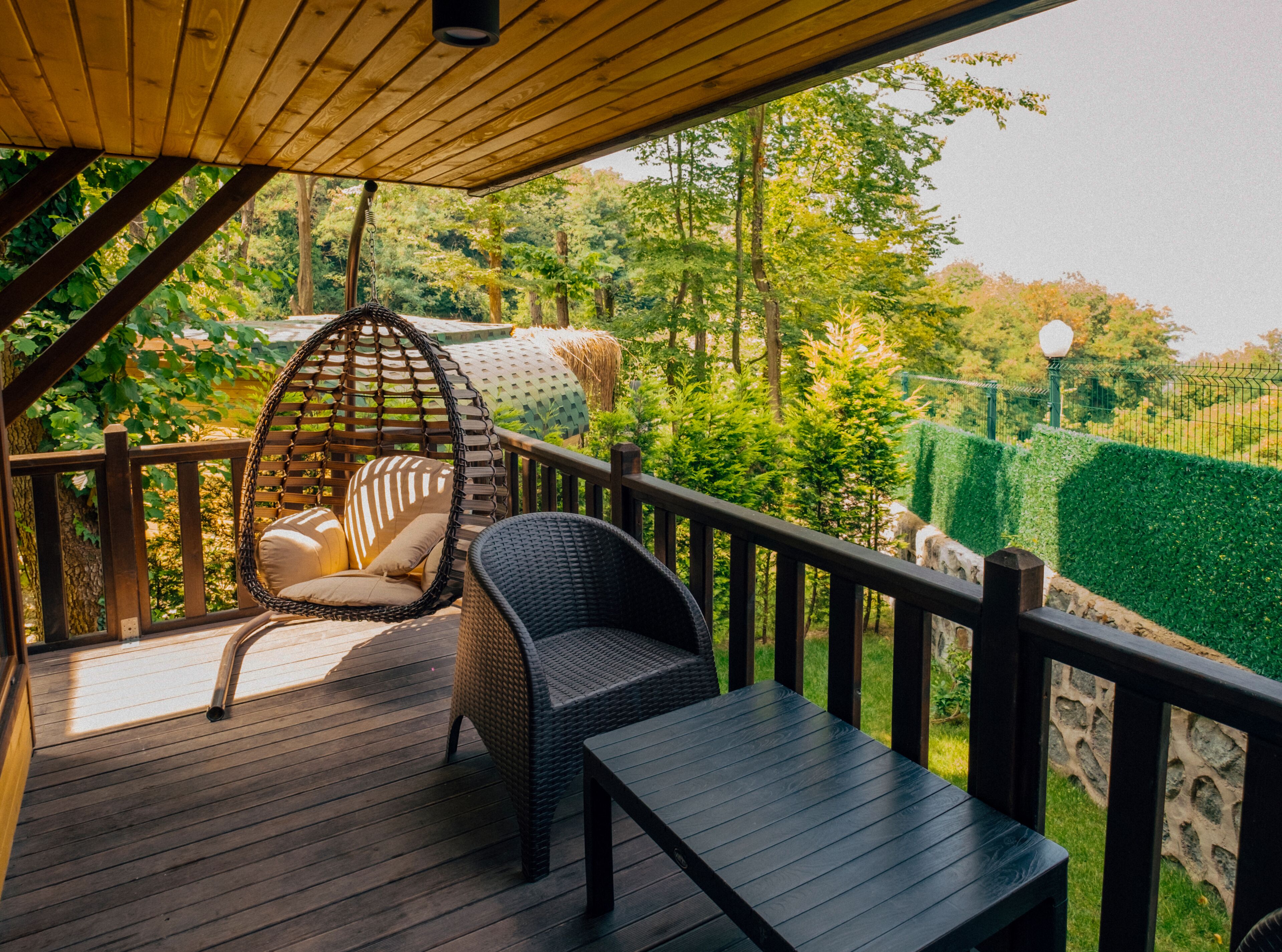 A wooden deck with a hanging chair and a bench, surrounded by greenery and a stone wall.