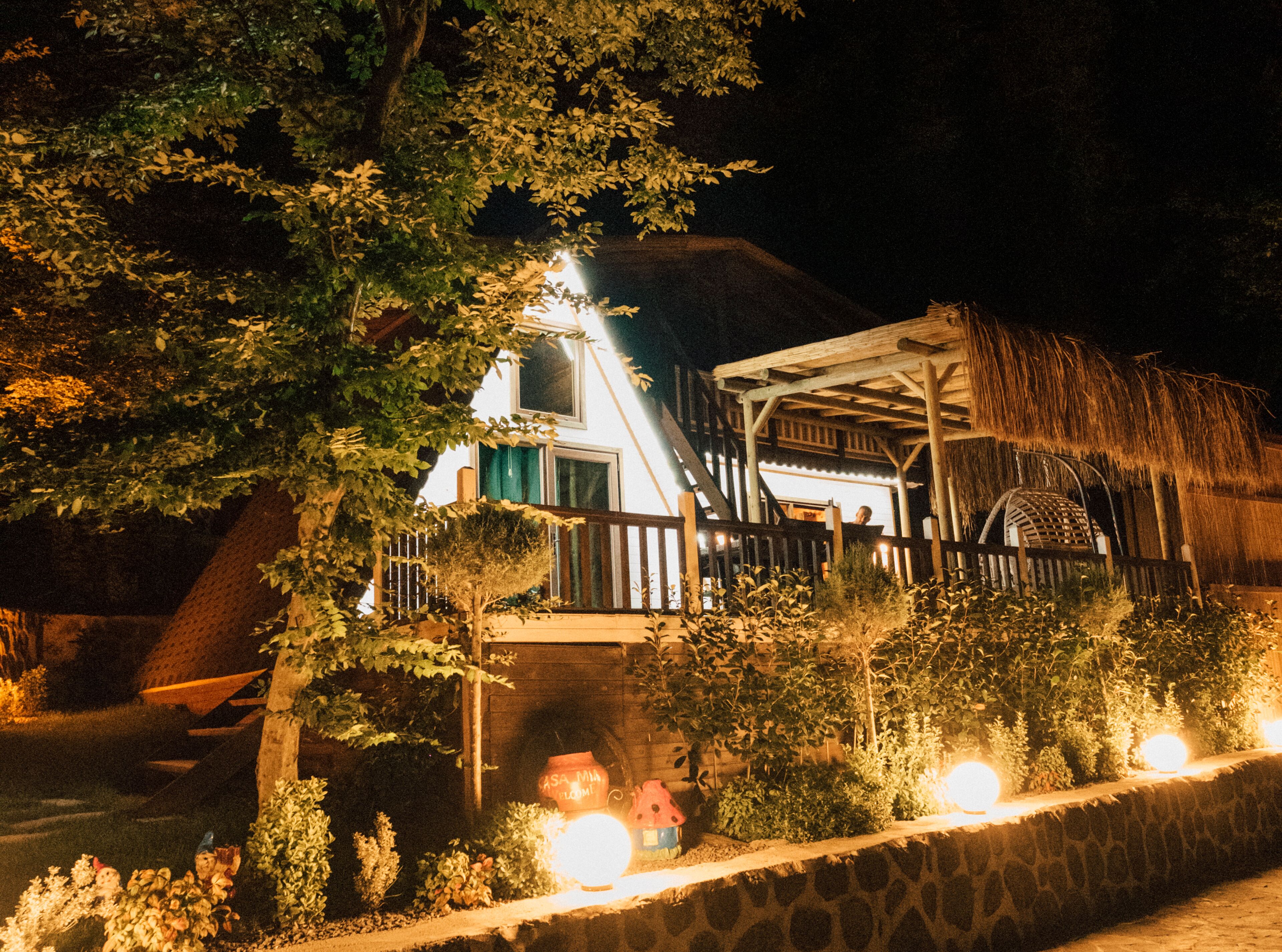A rustic building with a thatched roof, surrounded by lush greenery and illuminated by warm lights.