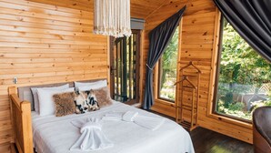 Une chambre dans une cabane en bois, avec un lit, une suspension en macramé, une fenêtre avec des rideaux et une chaise en bois.