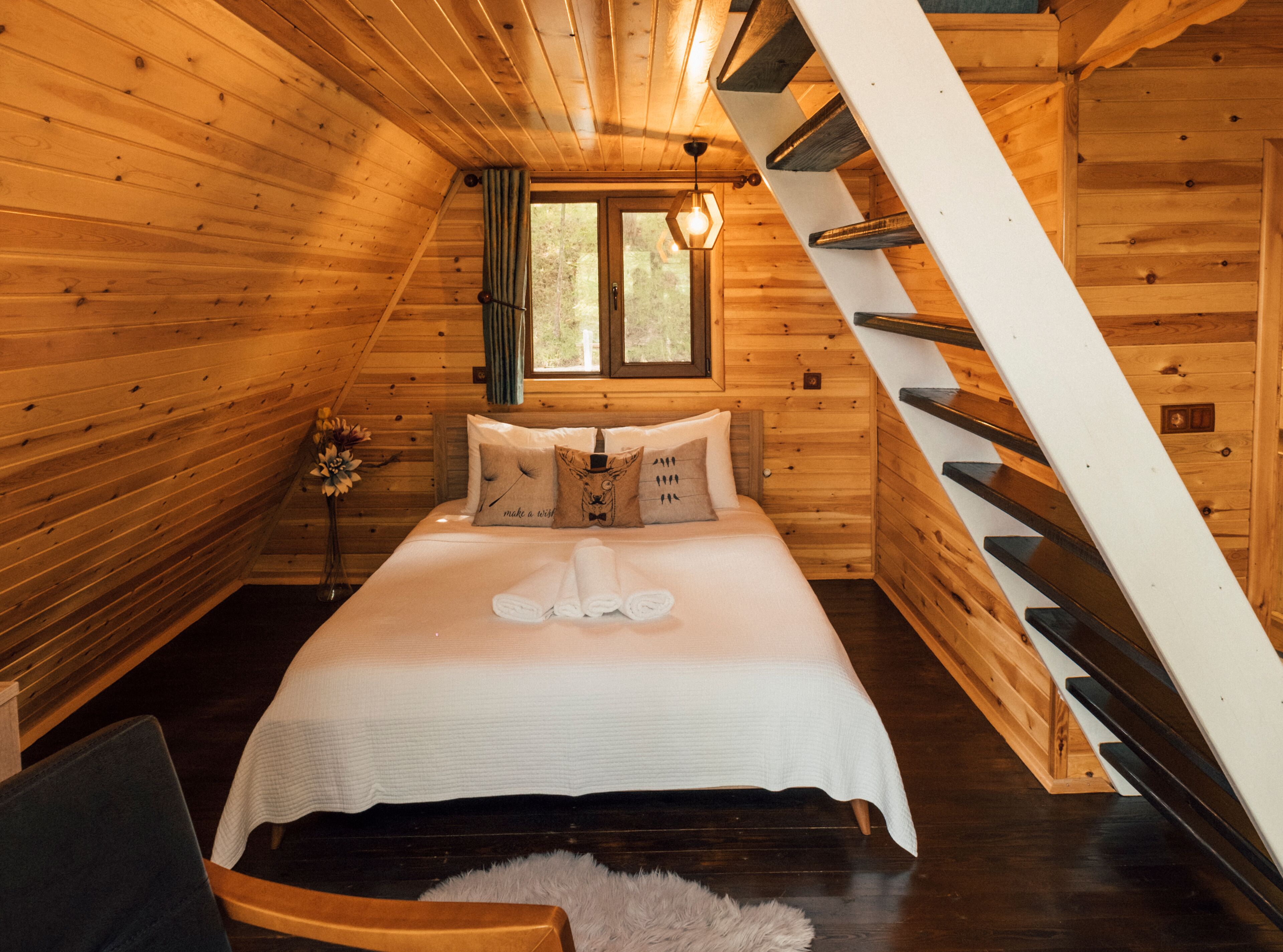 A cozy wooden cabin bedroom with a bed, pillows, a nightstand, a window, and a staircase.