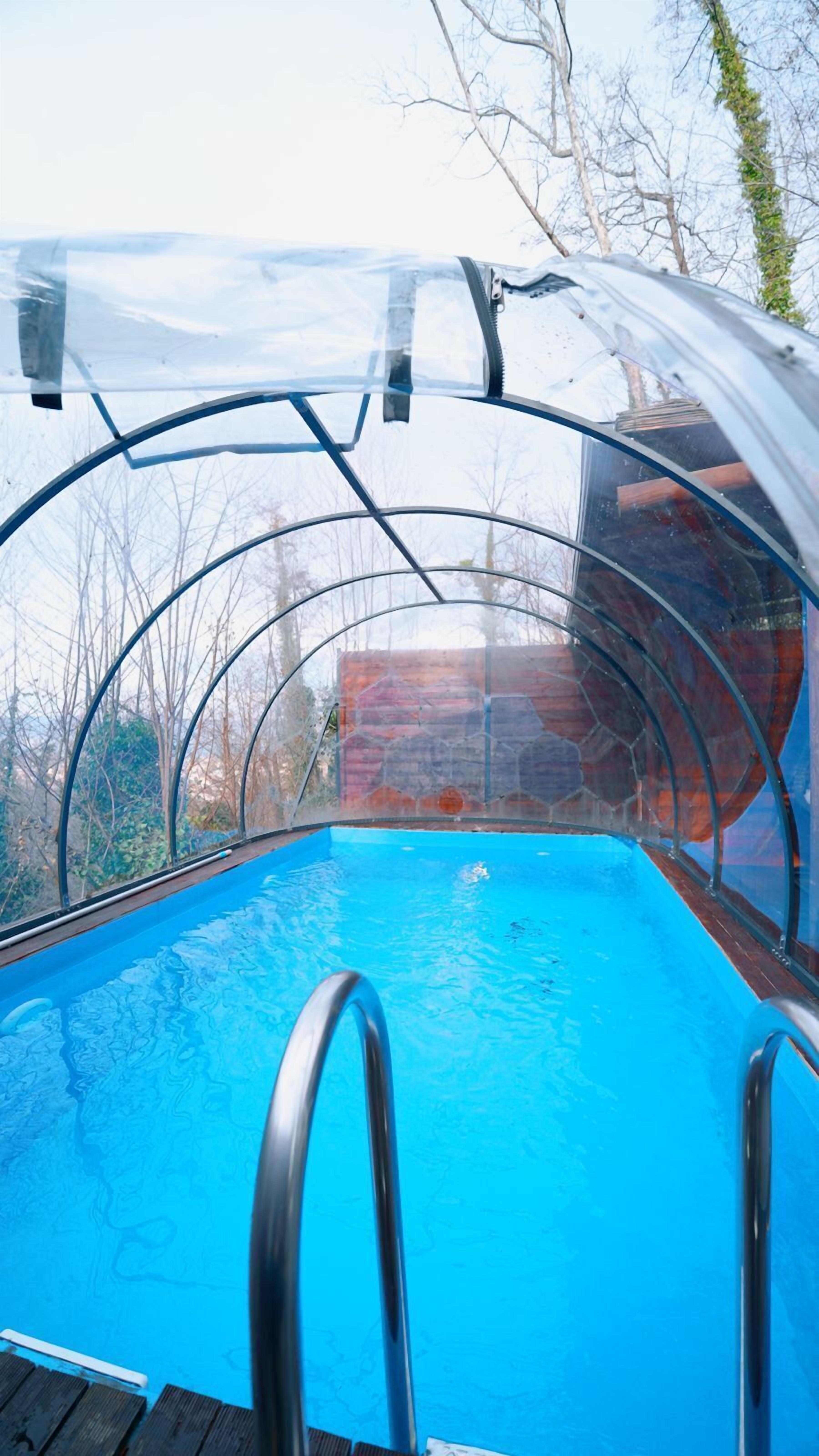 Una piscina transparente con barandillas de metal, rodeada de árboles y un muro de ladrillo.