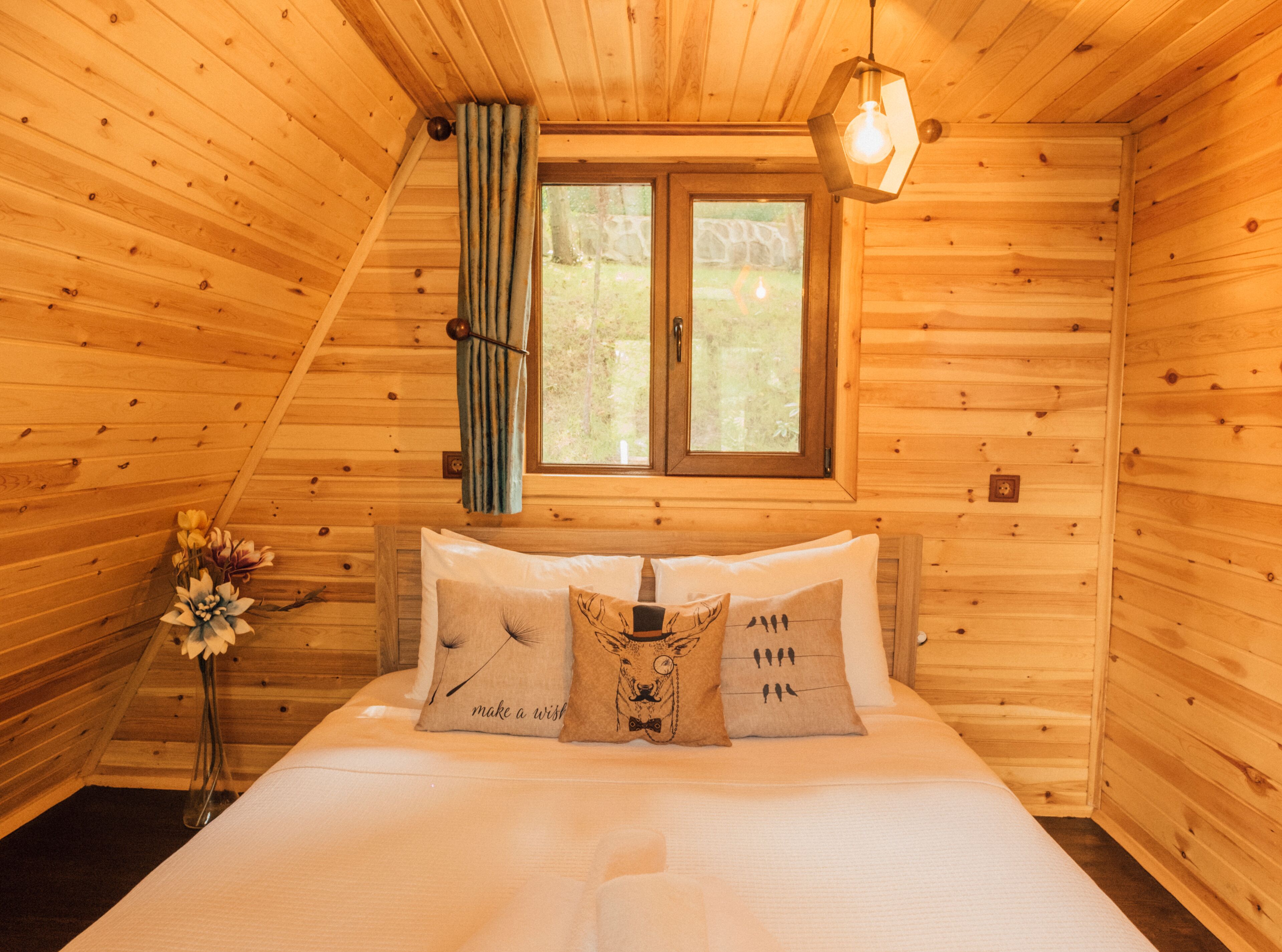 A wooden cabin bedroom with a bed, pillows, a window, and a hanging light fixture.
