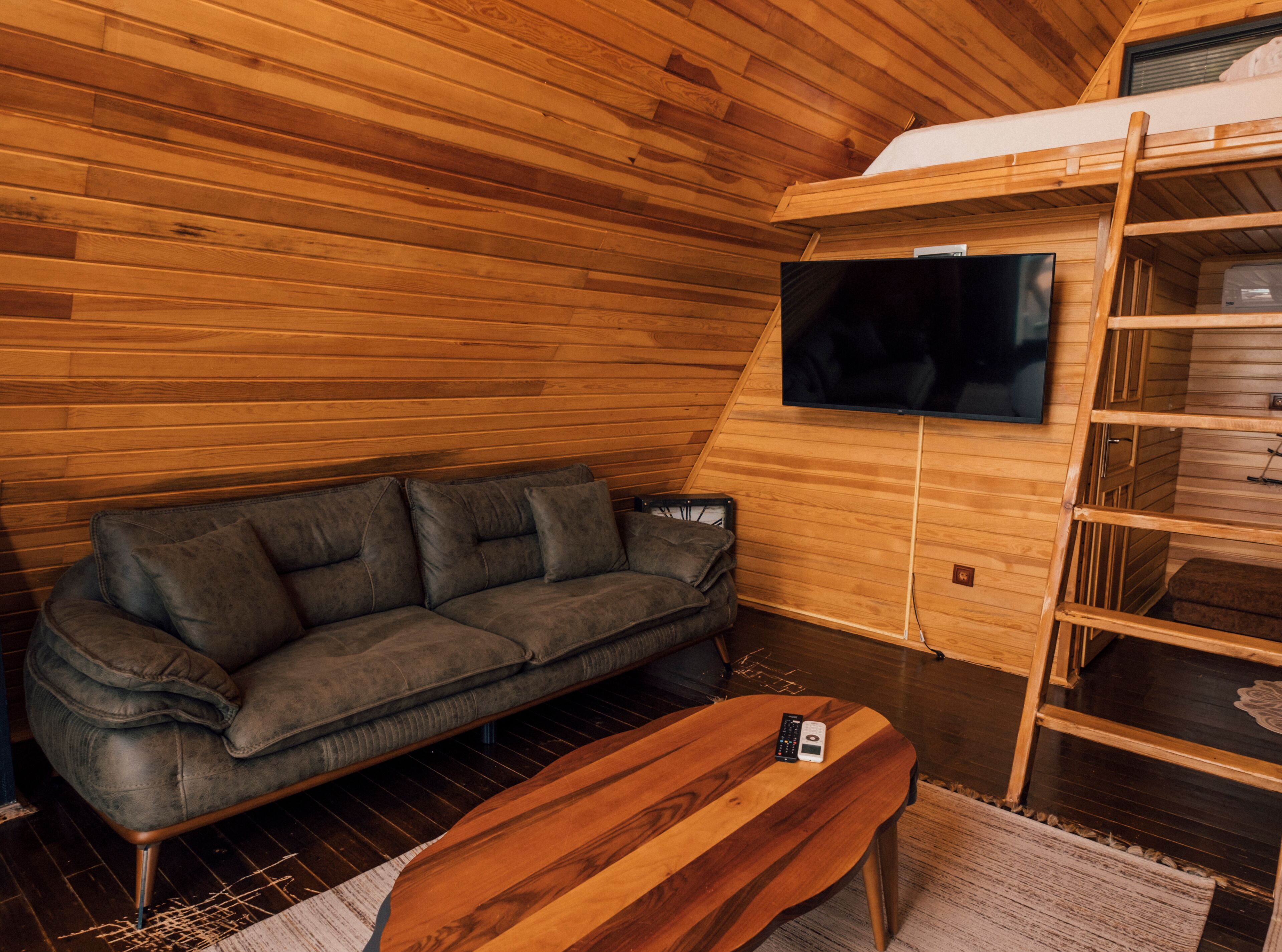 A cozy room with a wooden ceiling, a sofa, a TV mounted on the wall, and a bunk bed.
