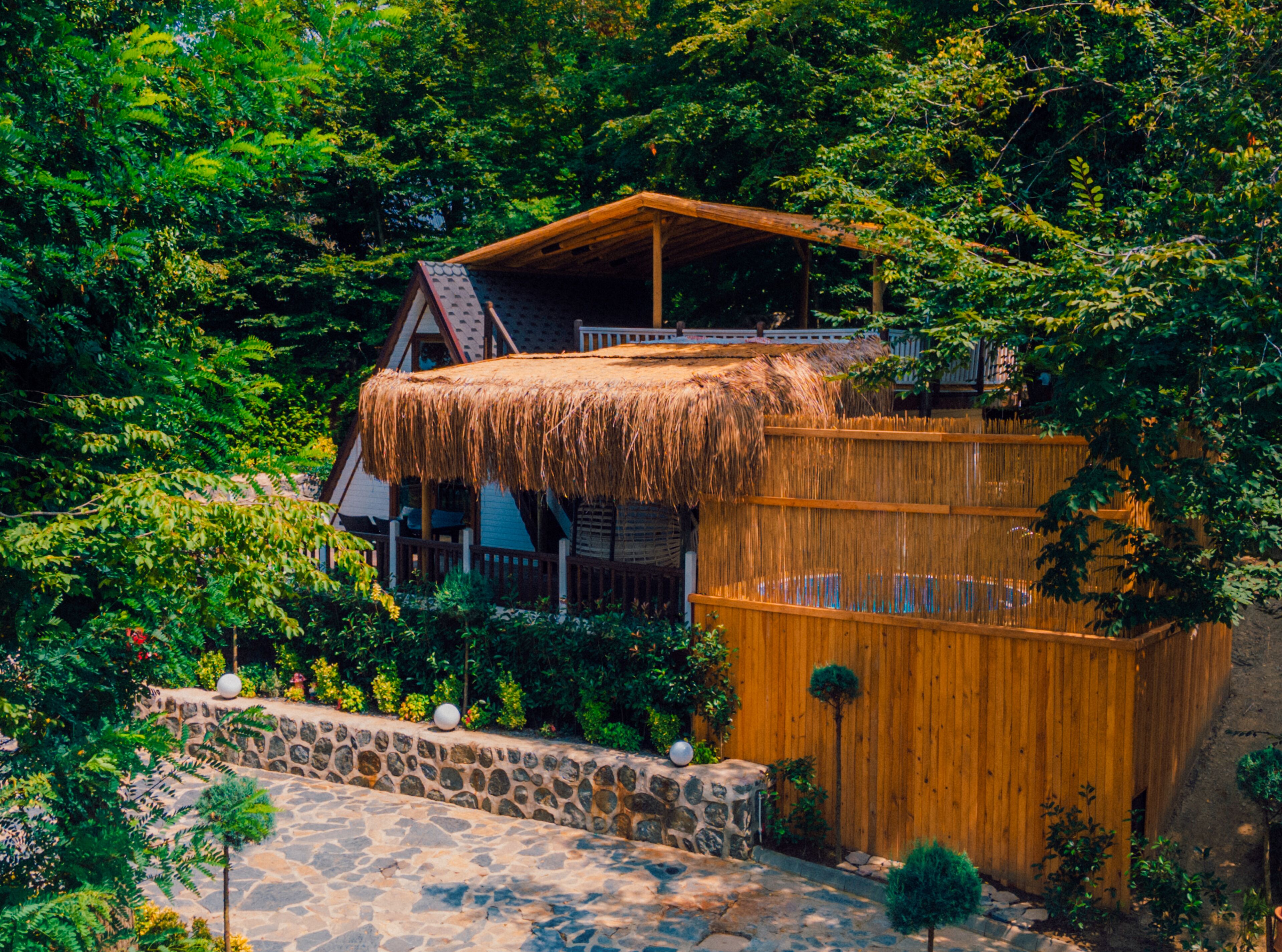 A wooden house with a thatched roof, surrounded by lush greenery and stone walls.