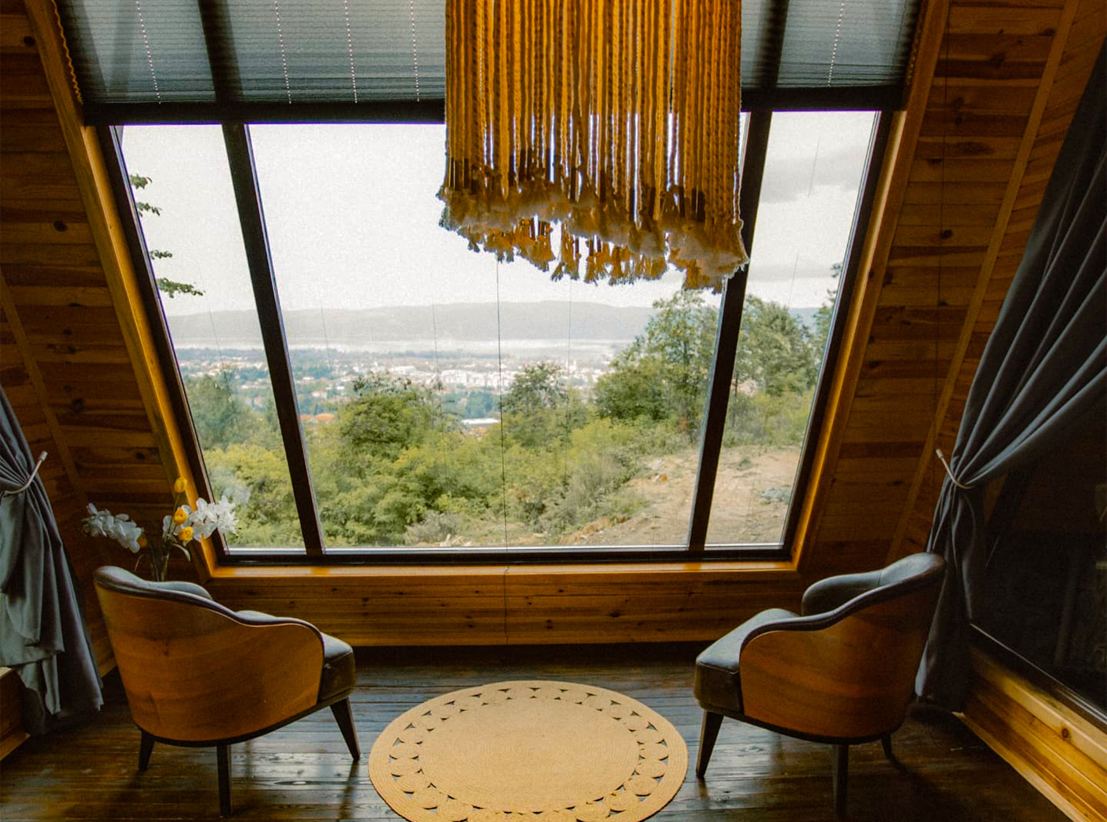 Una habitación con un ventanal grande, dos sillas, un tapiz tejido y una lámpara colgante.