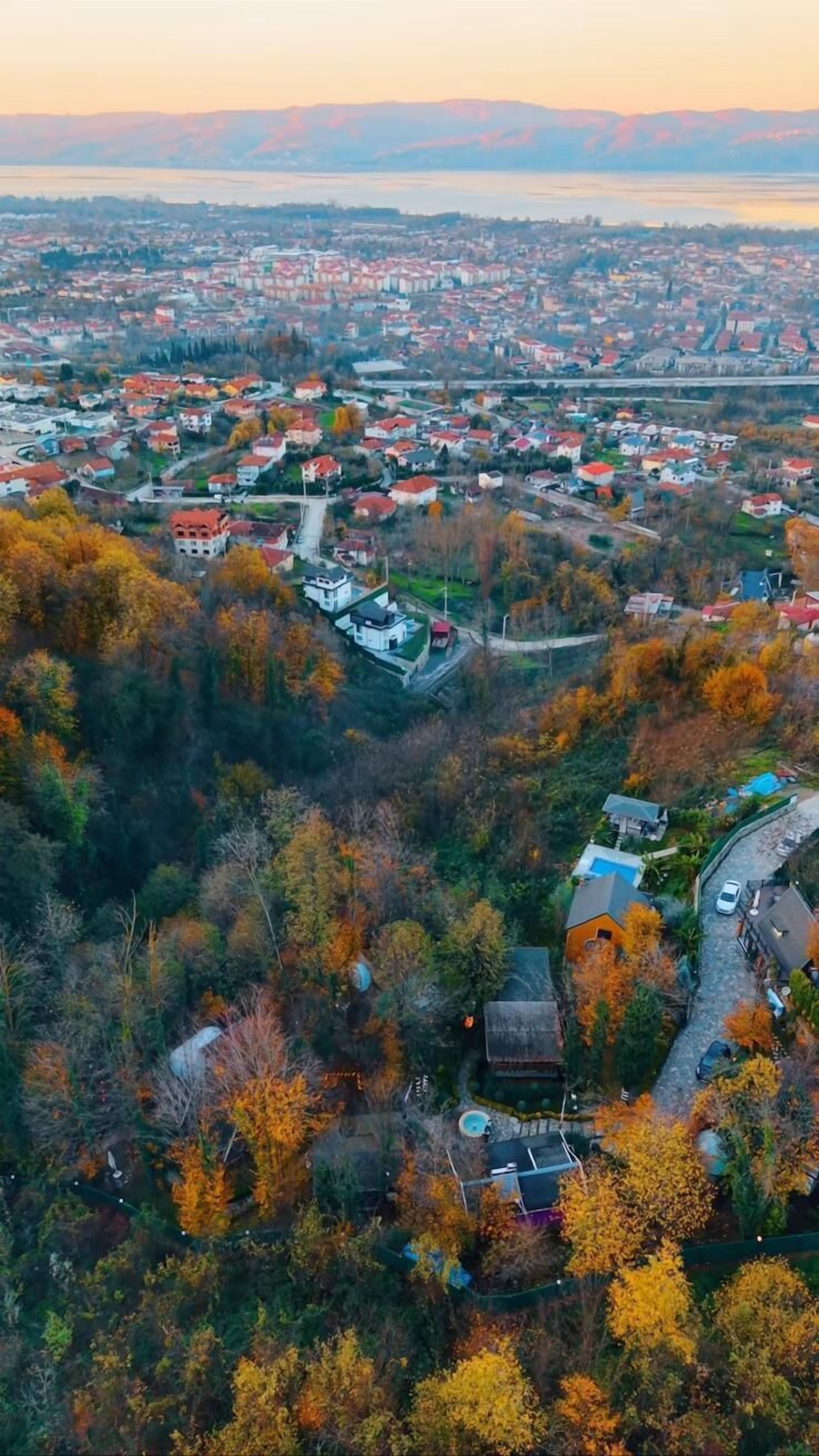 An aerial view of a town with a mix of residential buildings and natural landscapes, including autumn-colored trees and a body of water in the distance.