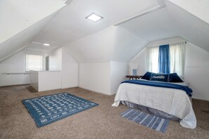 Ein Schlafzimmer mit schrägen Wänden, einem Bett mit blauem Kopfkissen, einem Fenster mit Vorhängen und einem blauen Teppich.