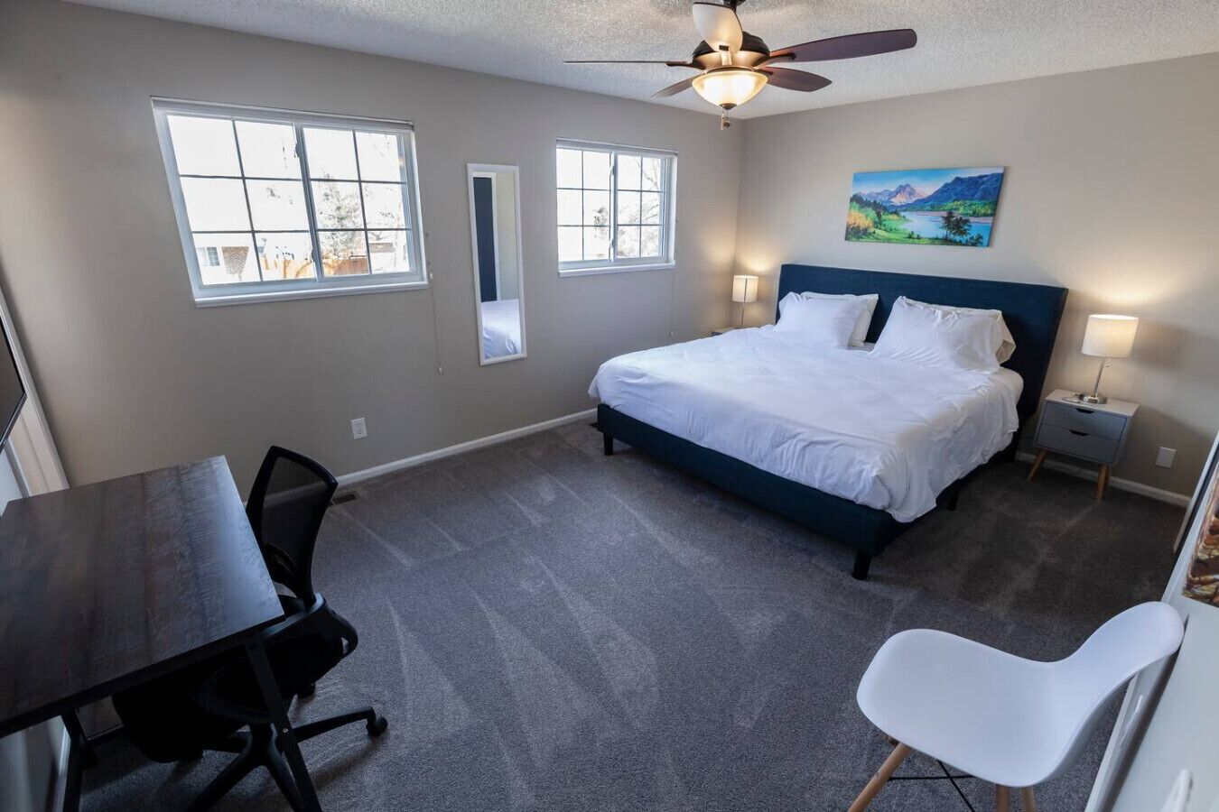 A bedroom with a bed, a desk, a chair, a ceiling fan, and two windows.