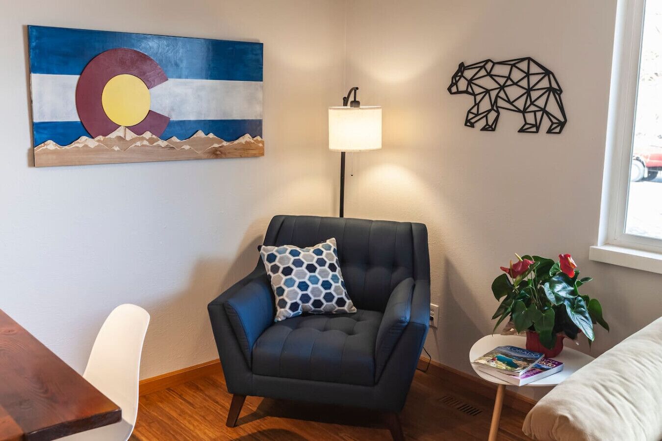 A room with a dark blue armchair, a wall-mounted lamp, a painting of a mountain landscape, a geometric wall art, and a potted plant.