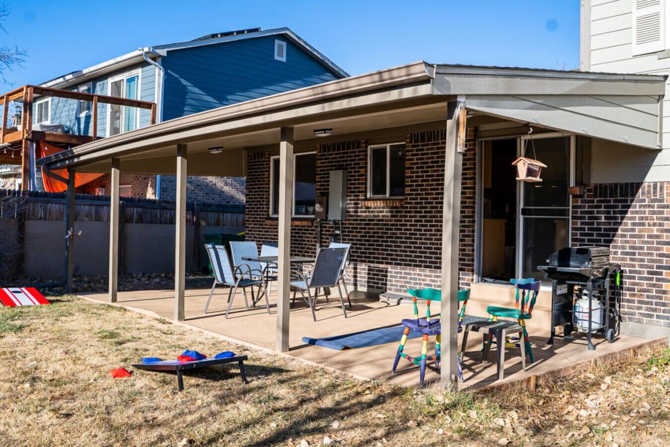 A covered patio with chairs and a table, a brick house, and a pool in the background.