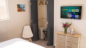 Ein Schlafzimmer mit einem Bett, einem Schrank mit Fernseher, einem Wäschekorb und einem kleinen Schreibtisch mit Blumenvase.