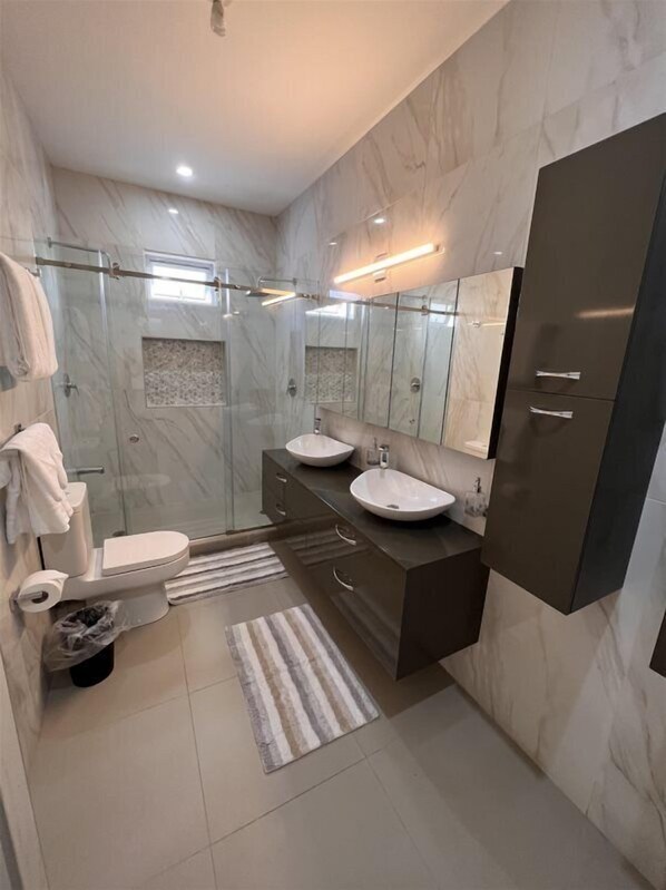 A modern bathroom with a glass shower enclosure, a toilet, two sinks, and a large mirror.