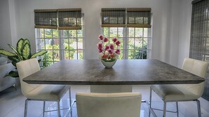 A modern dining area with a large rectangular table, two white leather chairs, and a bowl of pink flowers on the table. - Gulfview Townhouse With Pool and Jacuzzi (Diego Martin)