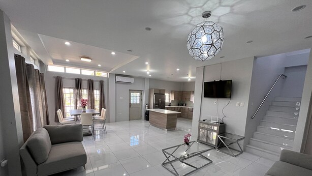 Una sala de estar moderna con un sofá, una mesa de centro de vidrio y un comedor con mesa y sillas. Hay una zona de cocina con un refrigerador y un televisor montado en la pared.