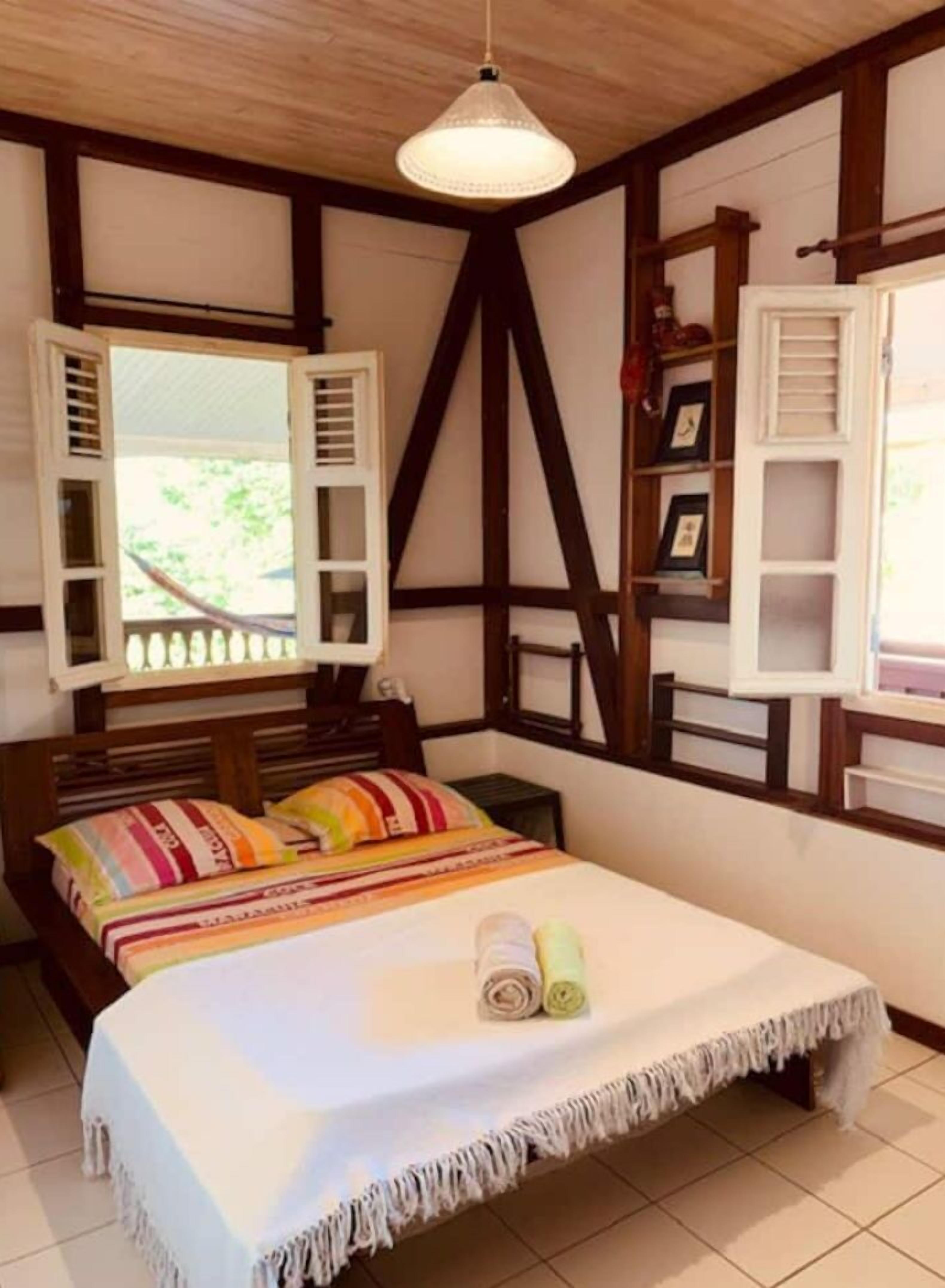 A bedroom with a bed, a window with shutters, a wooden ceiling, and a shelf with items.