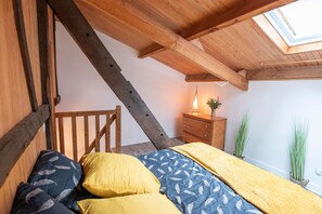 A loft bedroom with a sloped ceiling, wooden beams, a bed with patterned bedding, a dresser, and a skylight.