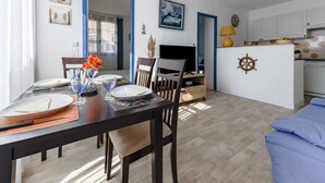 A dining area with a table set for two, a TV, and a kitchen in the background. - L'équinoxe - Charmant logement 150m de la plage (Leucate)