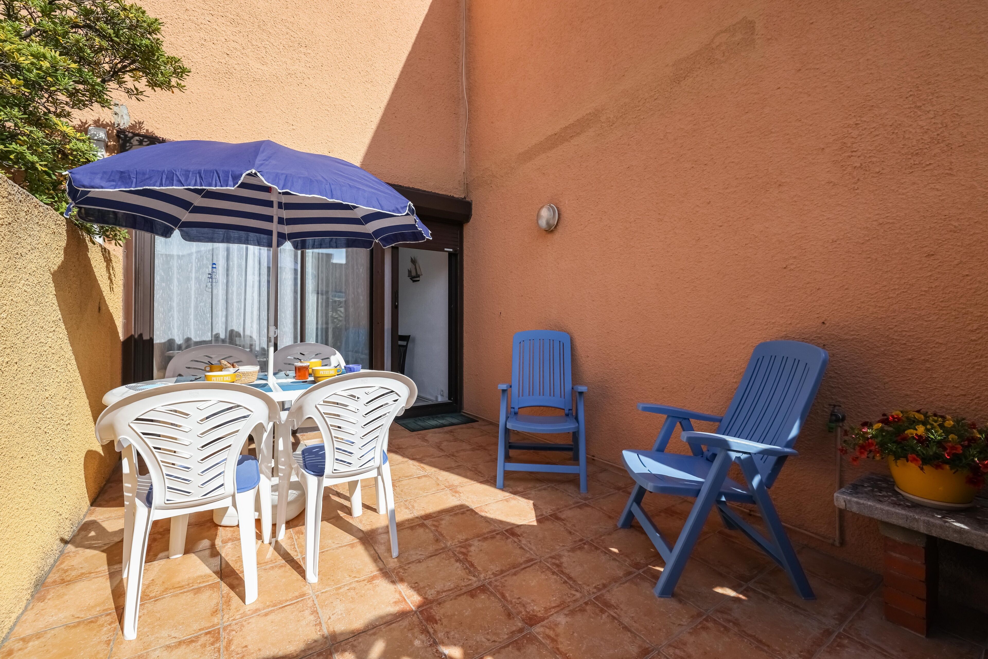 Een terras buiten met een tafel en stoelen, een blauwe parasol en een bloempot.