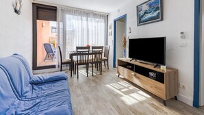 A living room with a blue sofa, a wooden TV stand, a dining table with chairs, and a large window with sheer curtains. - L'équinoxe - Charmant logement 150m de la plage (Leucate)