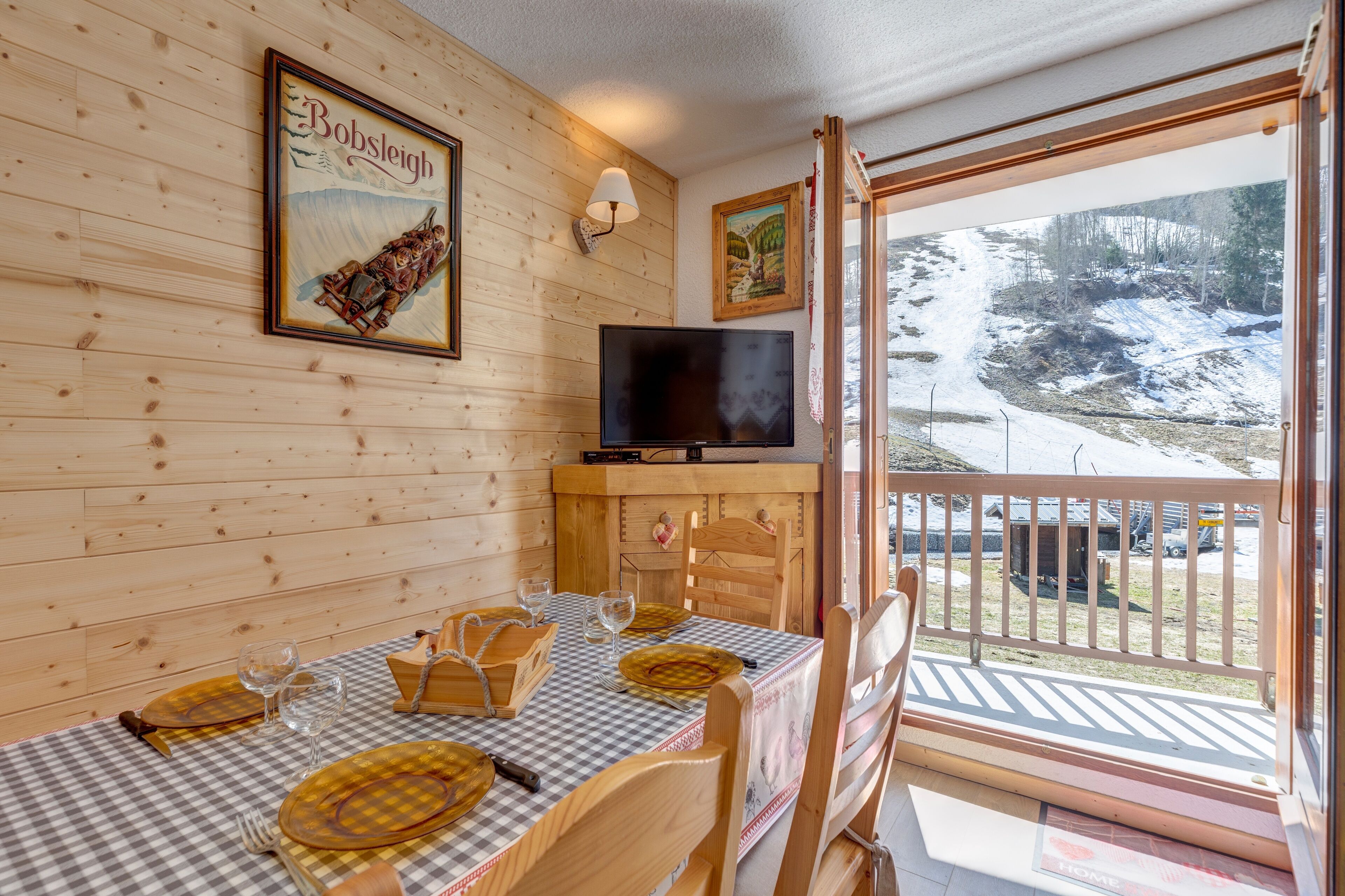 A wooden interior with a dining table set for two, a television on a stand, and a view of a snowy outdoor area.