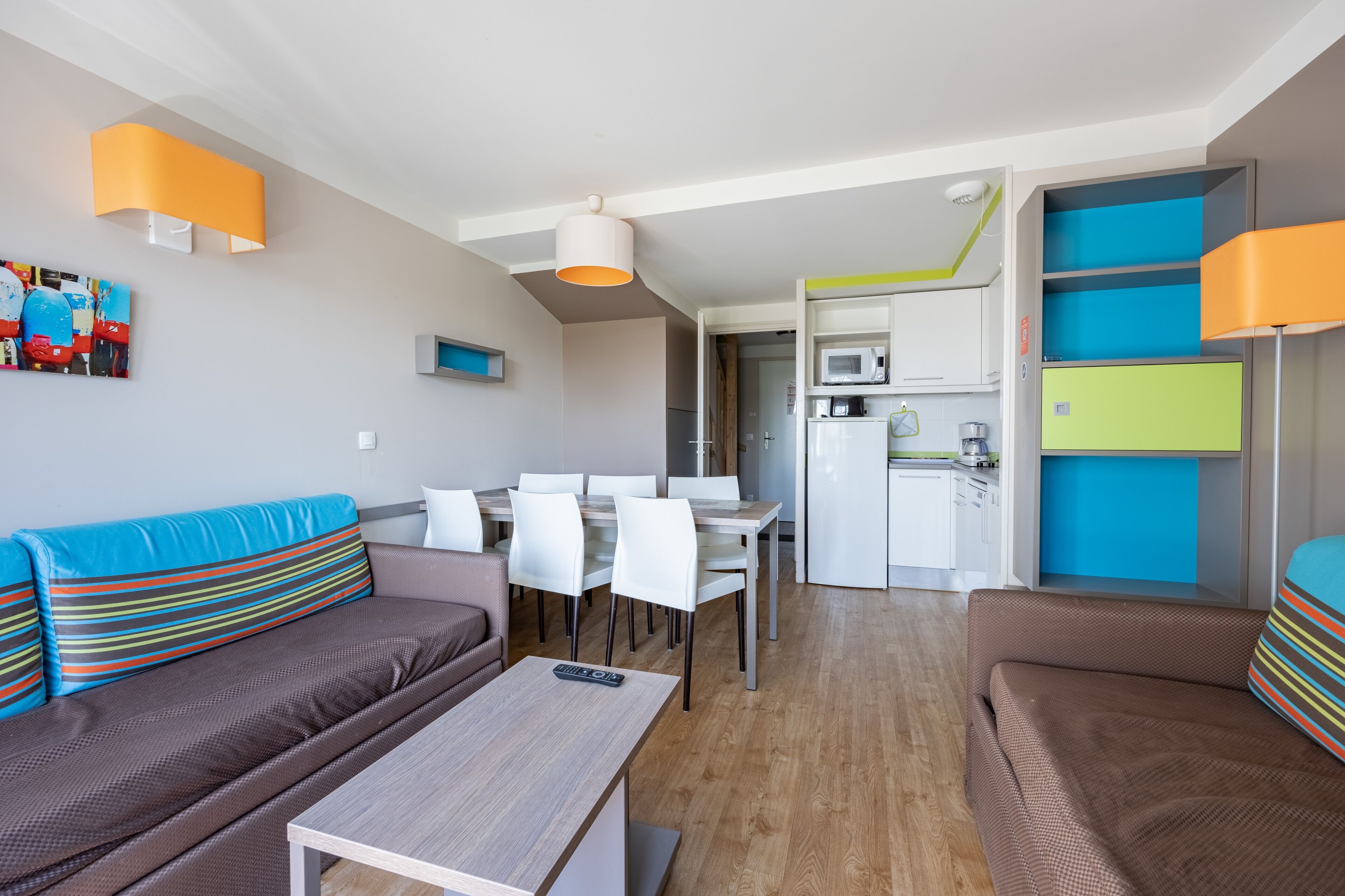 A modern living room with a sofa, dining table, and chairs. There is a kitchen area with a microwave and cabinets.