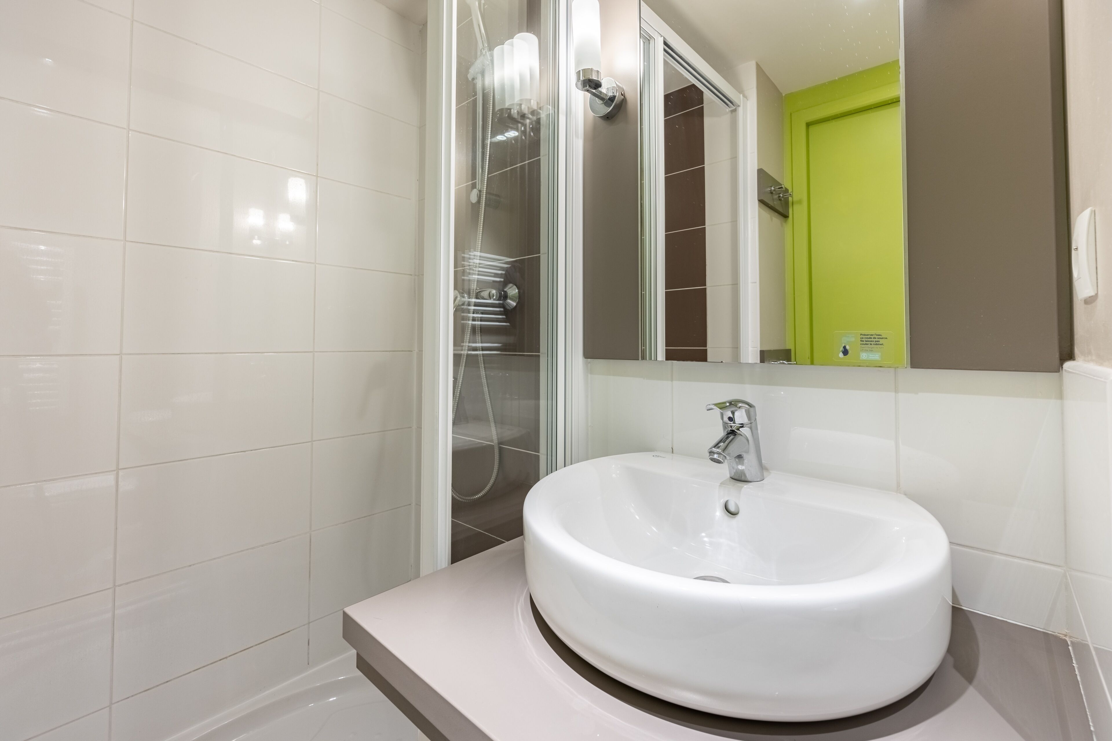 Une salle de bain moderne avec un lavabo blanc, une douche en verre et un miroir qui reflète une porte verte.