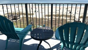 A balcony with two turquoise chairs and a small table, overlooking a beach with people and a wooden deck. - Blue Palmetto Vacation Rentals (Myrtle Beach)