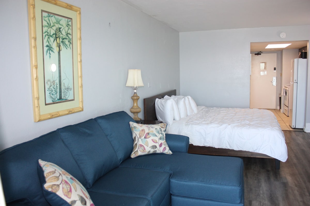 Une chambre à coucher avec un lit, un canapé bleu orné de coussins à motifs, une table de chevet avec une lampe et un miroir fixé au mur.