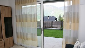 Een kamer met een glazen schuifdeur die uitkomt op een terras met uitzicht op een berg en een huis.