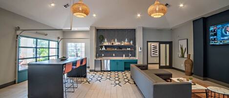 Une cuisine moderne avec un îlot central, un coin repas avec une table noire et des chaises orange, et un salon avec un canapé gris et une table basse.