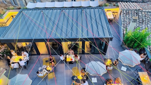 Un espace extérieur aménagé pour les repas, avec des tables et des chaises, un coin salon abrité et un bâtiment doté d’un toit en tuiles.