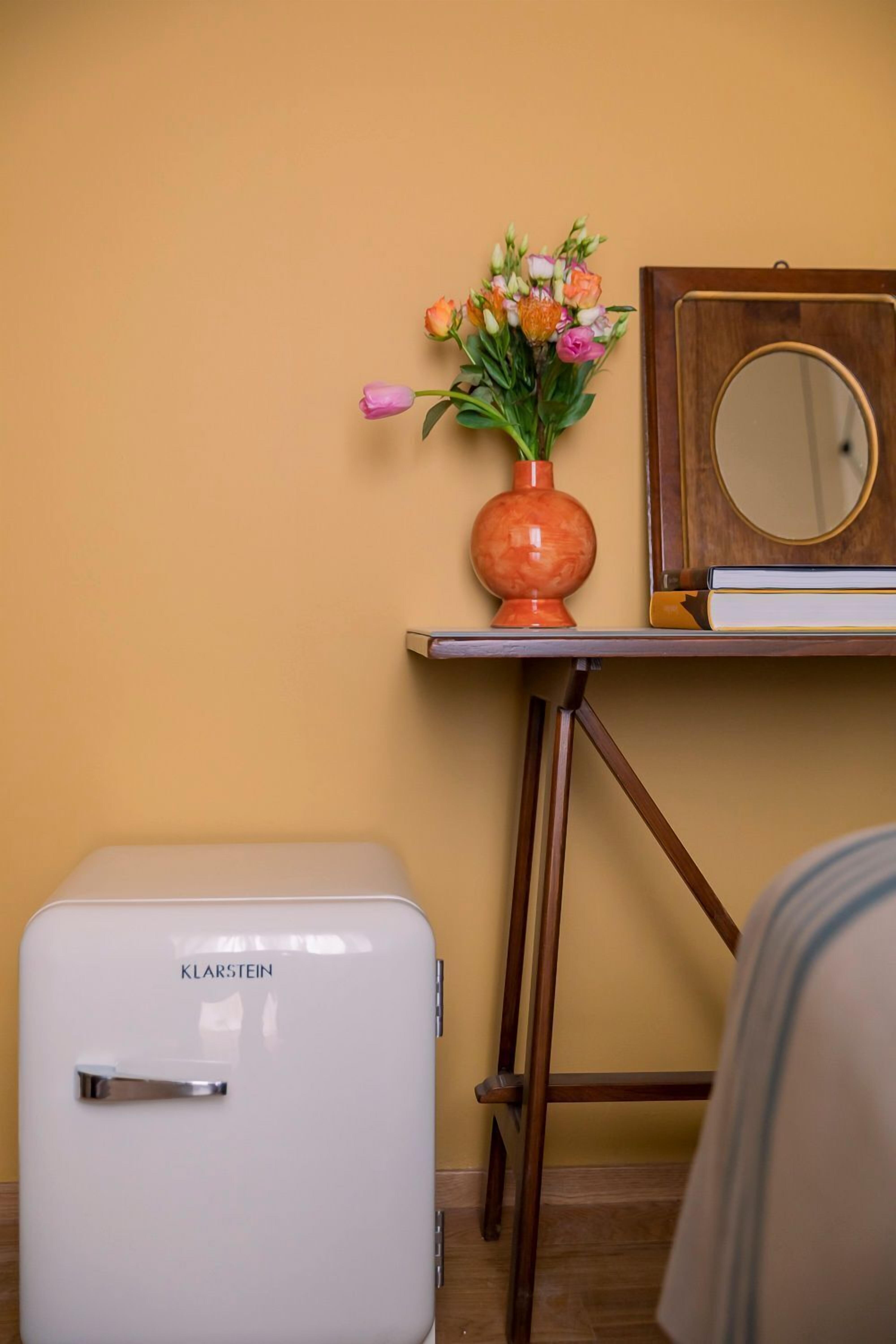 A small white refrigerator with the brand name 'Klarstein' on it, a wooden table with a mirror and a vase of flowers, and a wooden floor.