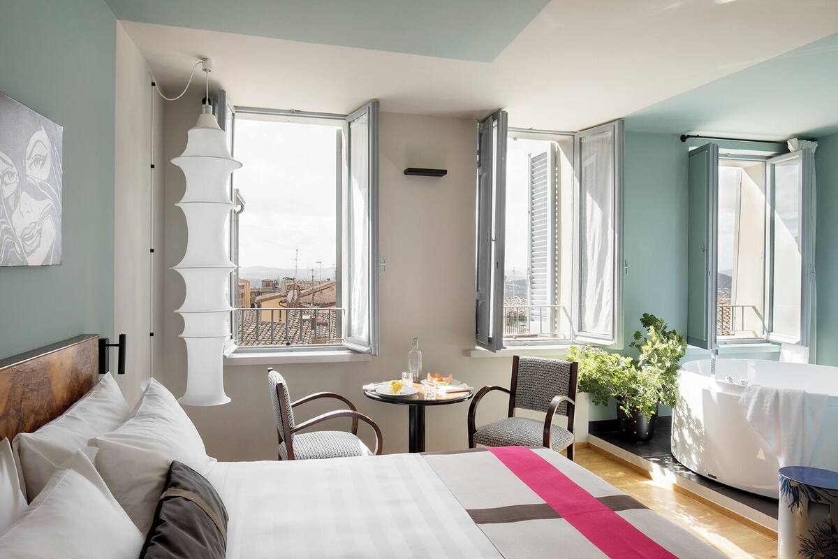 A hotel room with a bed, a small table with a chair, a window with a view of buildings, and a bathroom with a bathtub.