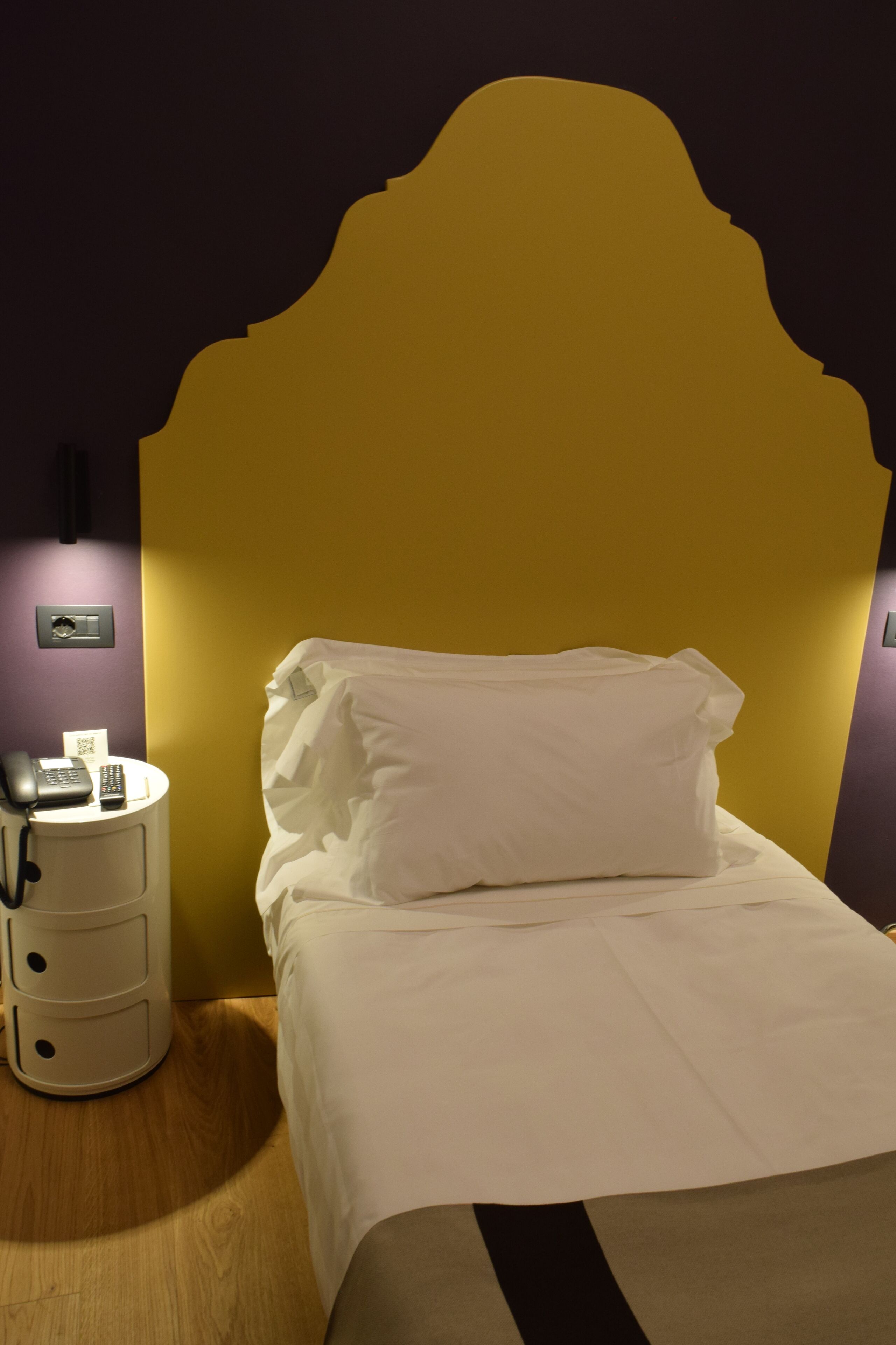 A neatly made bed with white linens and pillows, a bedside table with a phone and remote control, and a wall-mounted light fixture.