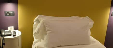 A neatly made bed with white linens and pillows, a bedside table with a phone and remote control, and a wall-mounted light fixture.