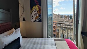 Une chambre d’hôtel avec un lit, un tableau, une fenêtre donnant sur des immeubles et un balcon.