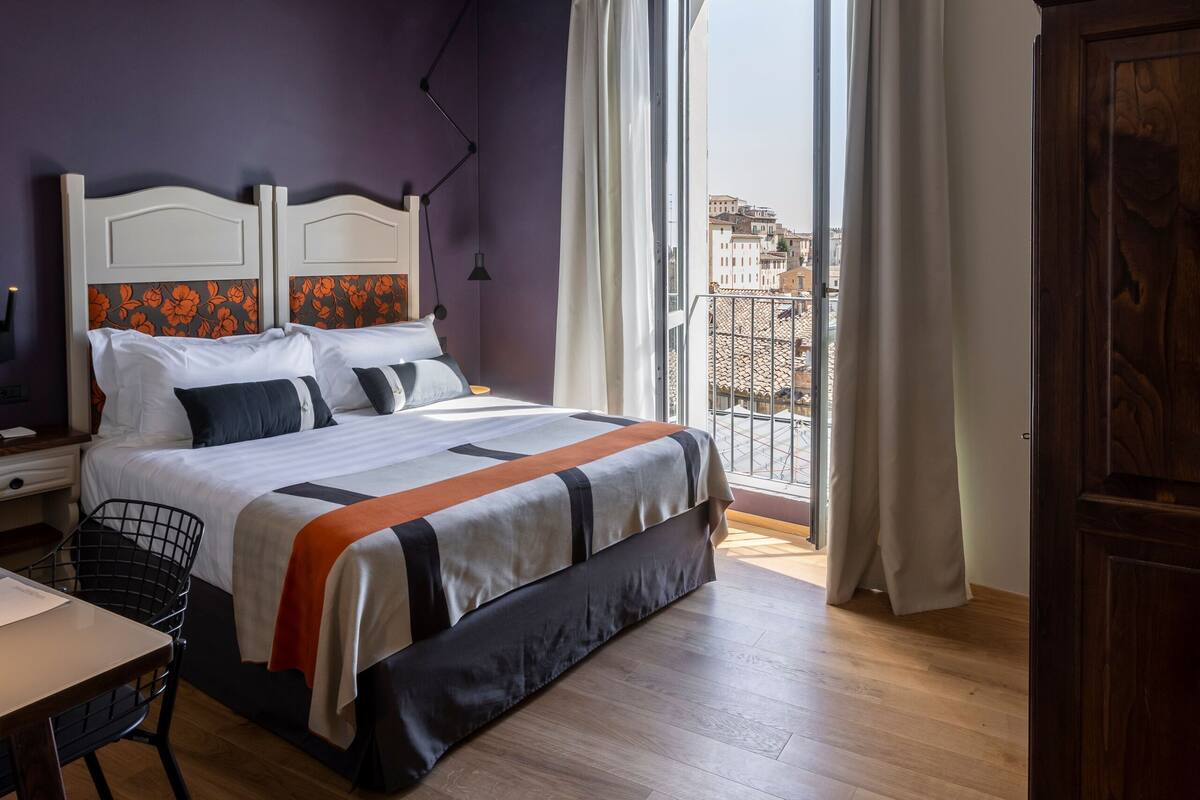 A bedroom with a bed, a desk, a chair, a wardrobe, and a view of buildings through a window.
