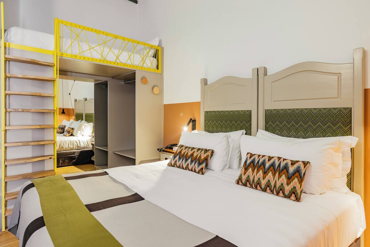A hotel room with a bunk bed, a desk with a lamp, and a bed with patterned pillows.