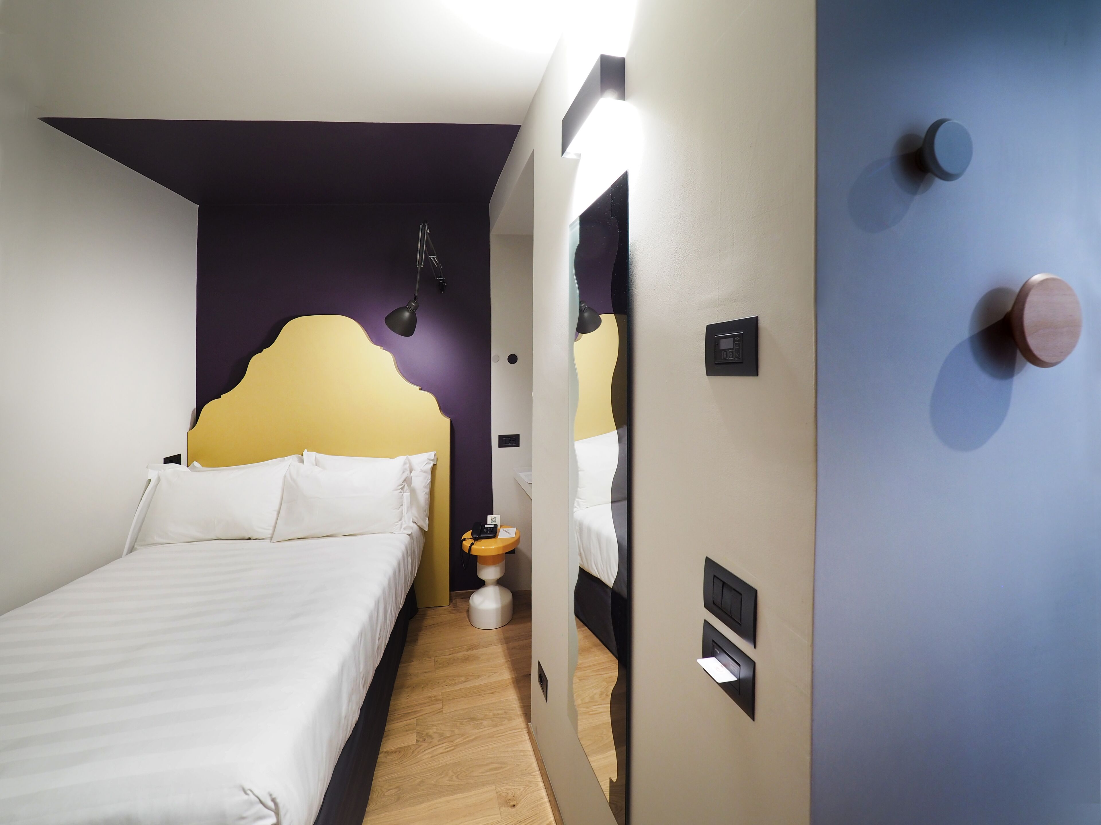 A modern hotel room with a bed, bedside table, lamp, and a mirror with a wooden door.