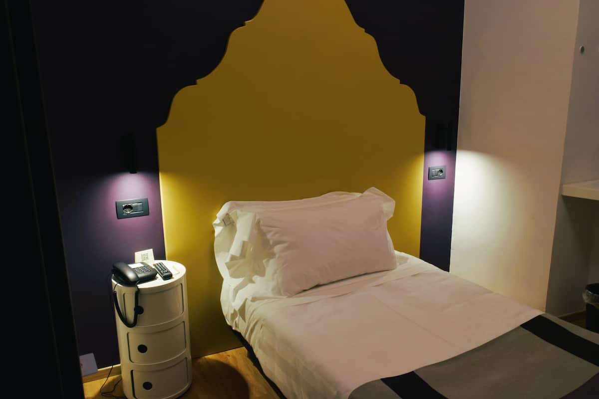 A bed with white bedding and a headboard, a bedside table with a phone and remote, and wall-mounted light switches.
