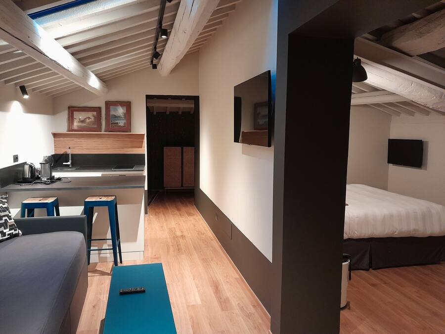 A compact hotel room with a kitchenette, a bed, a seating area with a blue mat, and a TV mounted on the wall.