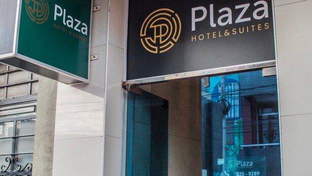 Entrada de un hotel con un letrero que dice "Plaza Hotel & Suites" y una vista parcial de un coche a través del vidrio.