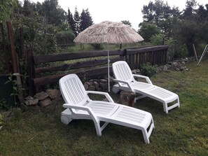Two white lounge chairs with a thatched umbrella in a grassy outdoor area, surrounded by a wooden fence and trees.