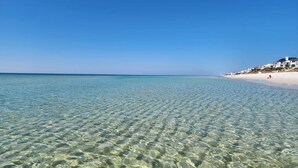 A clear beach with shallow water, a sandy shore, and a row of white buildings along the coastline. - Beach Bliss: Beach Minutes Away, 6-Seater Golf Cart, and Sparkling Resort Pool! (Santa Rosa Beach)