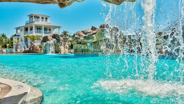 A swimming pool with clear turquoise water, a waterfall feature, and a large building in the background. - Beach Bliss: Beach Minutes Away, 6-Seater Golf Cart, and Sparkling Resort Pool! (Santa Rosa Beach)