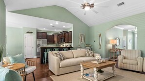 A spacious living room with a sofa, coffee table, and chairs. A kitchen with dark cabinets and a dining area with a table and chairs are visible in the background. - Beach Bliss: Beach Minutes Away, 6-Seater Golf Cart, and Sparkling Resort Pool! (Santa Rosa Beach)
