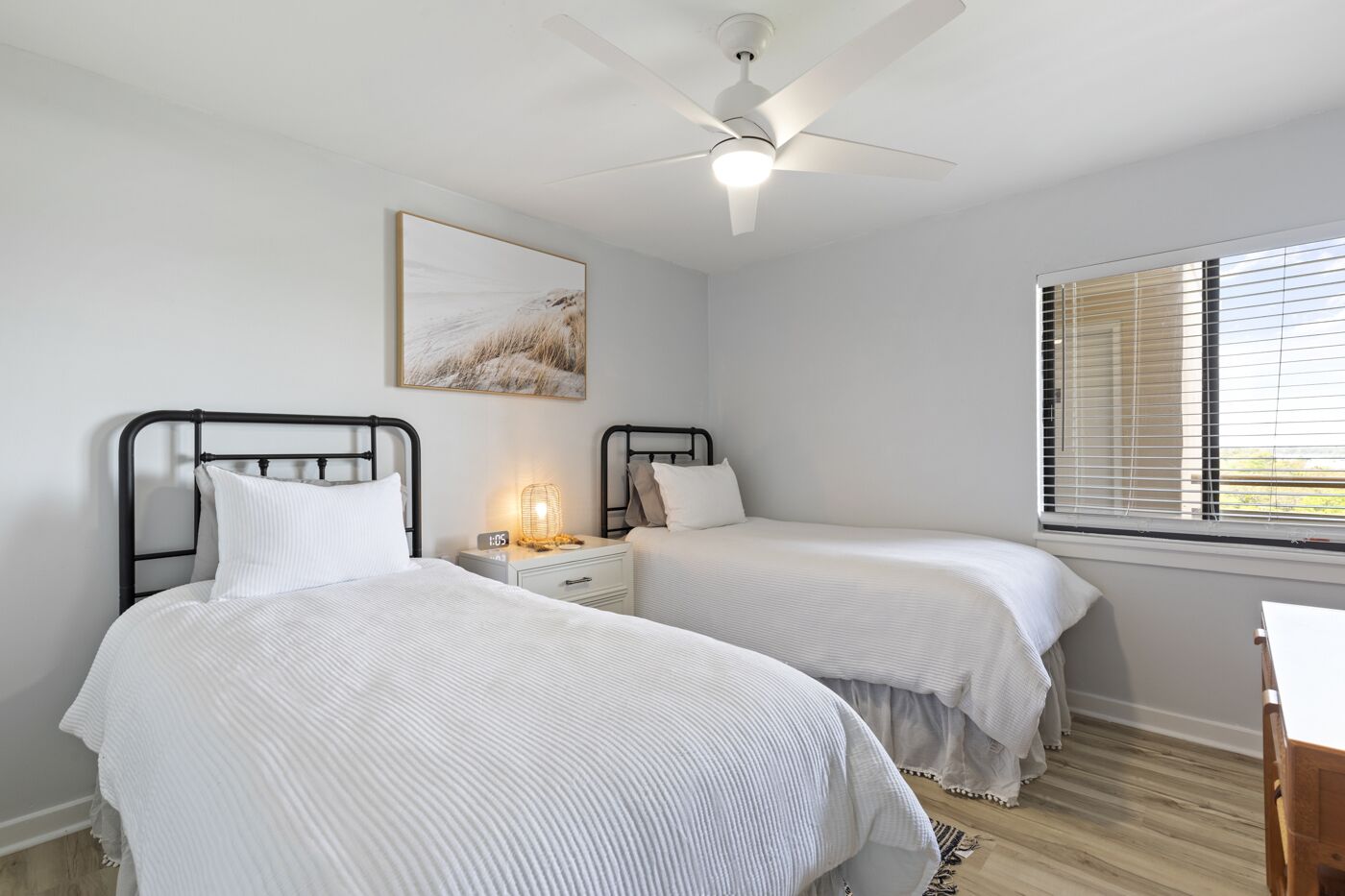 A bedroom with two beds, a ceiling fan, a bedside table with a lamp, and a window with blinds.