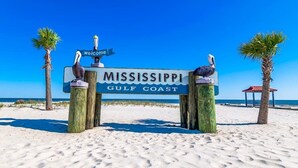 Un panneau en bois portant l’inscription « MISSISSIPPI GULF COAST » et une banderole « bienvenue », flanqué de deux poteaux en bois surmontés de sculptures d’oiseaux, le tout sur fond de plage.