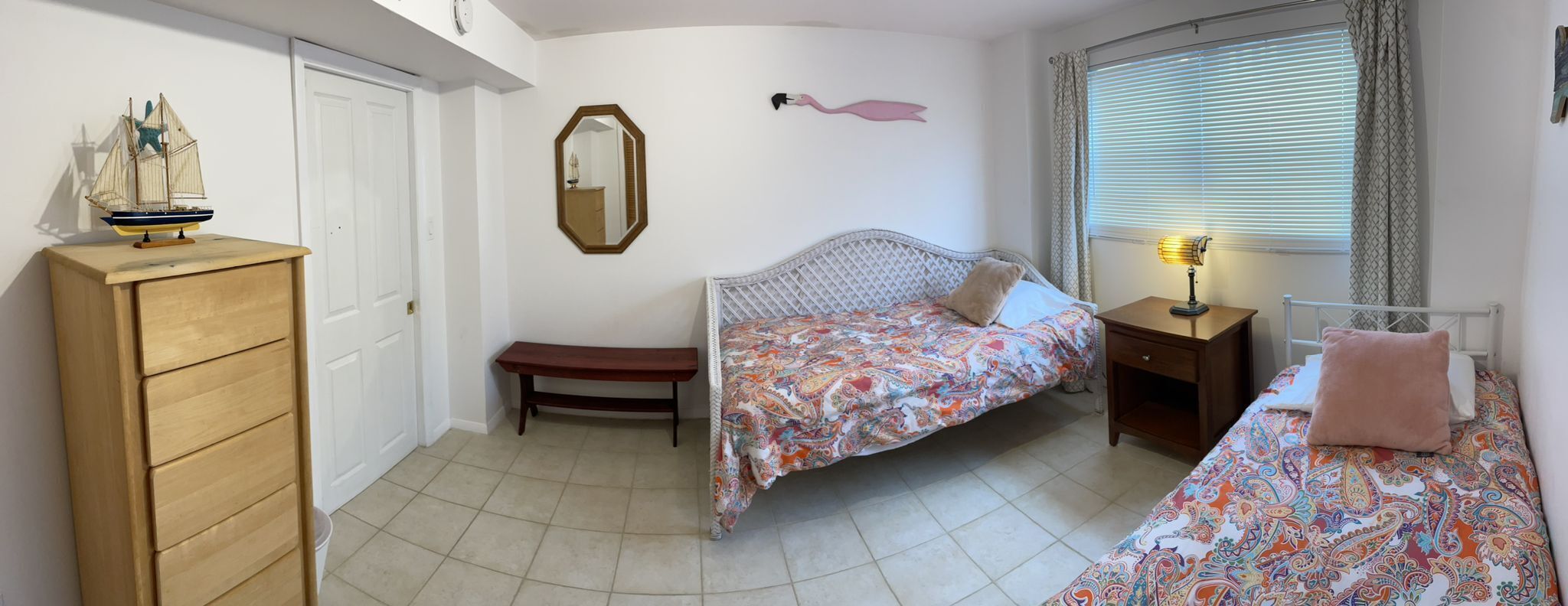 A bedroom with two beds, a wooden dresser, a bench, a mirror, and a window with blinds.