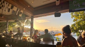 Un bar con vista sul tramonto sull'acqua, palme e alcuni tavoli occupati.